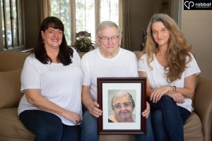 80 year old man in middle with his grown daughters on either side of him. They are all wearing white t-shirt. He is holding a photo of his deceased wife.