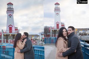Woman on left and man on right wearing winter coats in front of the white lighthouse in Port Credit with a red door and red roof. Two side by side vertical photos. They are looking at the camera in the photo on the right and kissing in the photo on the left.