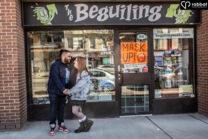Couple in front of Beguiling Books storefront. They are holding hands toward the left third of the photo. Woman is wearing a jean jacket, short skirt and black boots with white socks. Her legs are bare. Man is wearing a jean jacket, T-shirt and jeans. Man is Black and woman Metis and has a light complexion with red hair.