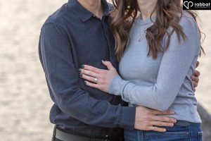 Close-up photo from the waist to the neck. Man is wearing a dark blue shirt and woman is wearing a longsleeve blueish gray shirt. They are hugging and you can see one of the man's hands and the woman's hand with her ring on it. There is water in the background.