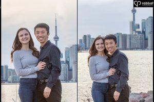 Cherry Beach engagement session. Vertical photos of a couple with the Toronto skyline including Lake Ontario and the CN Tower in the background. They are smiling at the camera. woman is wearing a long sleeve grayish blue shirt and the man is wearing a dark shirt. The man is Asian and the woman is white with brown hair.