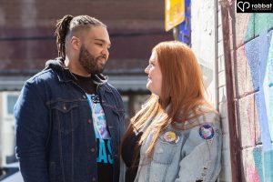 Backlit couple in front of an artistic mural looking lovingly at each other. Woman is wearing a jean jacket. Man is wearing a jean jacket with a T-shirt underneath. Man is black and woman Metis with has a light complexion and red hair.