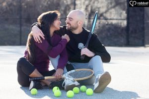 Sporty engagement photo. Couple sitting on tennis court. They are each holding wooden tennis courts. They have lots of tennis balls in front of them. They are looking at each other and smiling. Woman is South Asian, wearing a purple shirt and black leggings. Man is white, wearing a black long sleeved shirt and blue jeans.