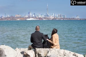 Couple in front of Lake Ontario sitting on rocks with their dog in-between them and the Toronto skyline in the background. The water is very blue.